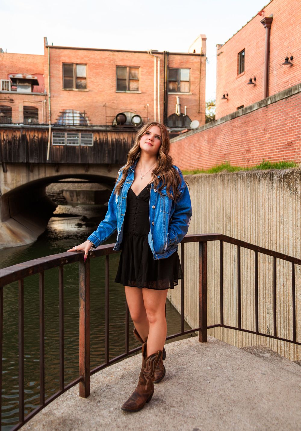Young woman in denim jacket, black dress, and cowboy boots posing on urban bridge walkway near river.