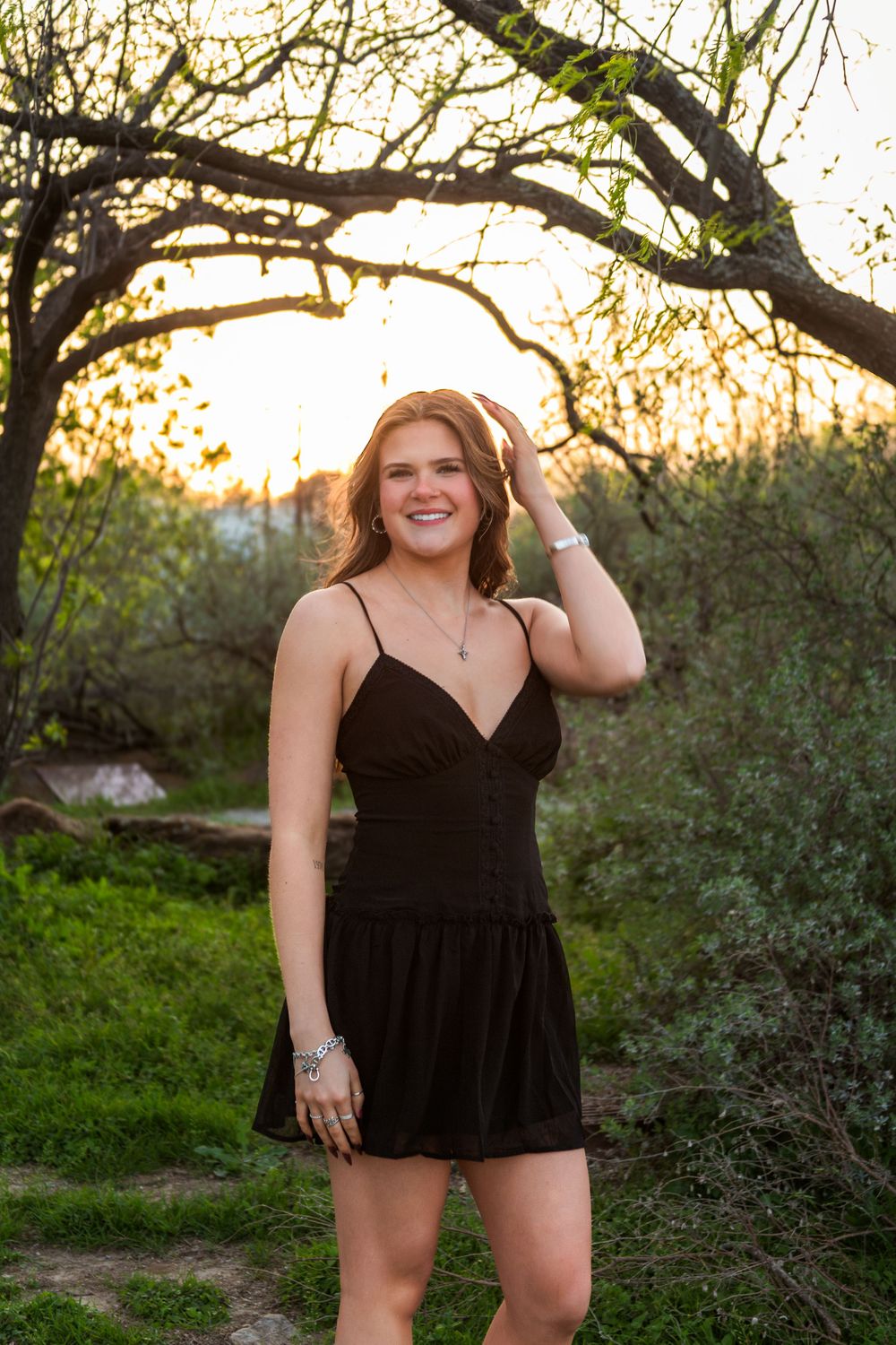 Young woman in black sundress smiling outdoors at golden hour among green trees.