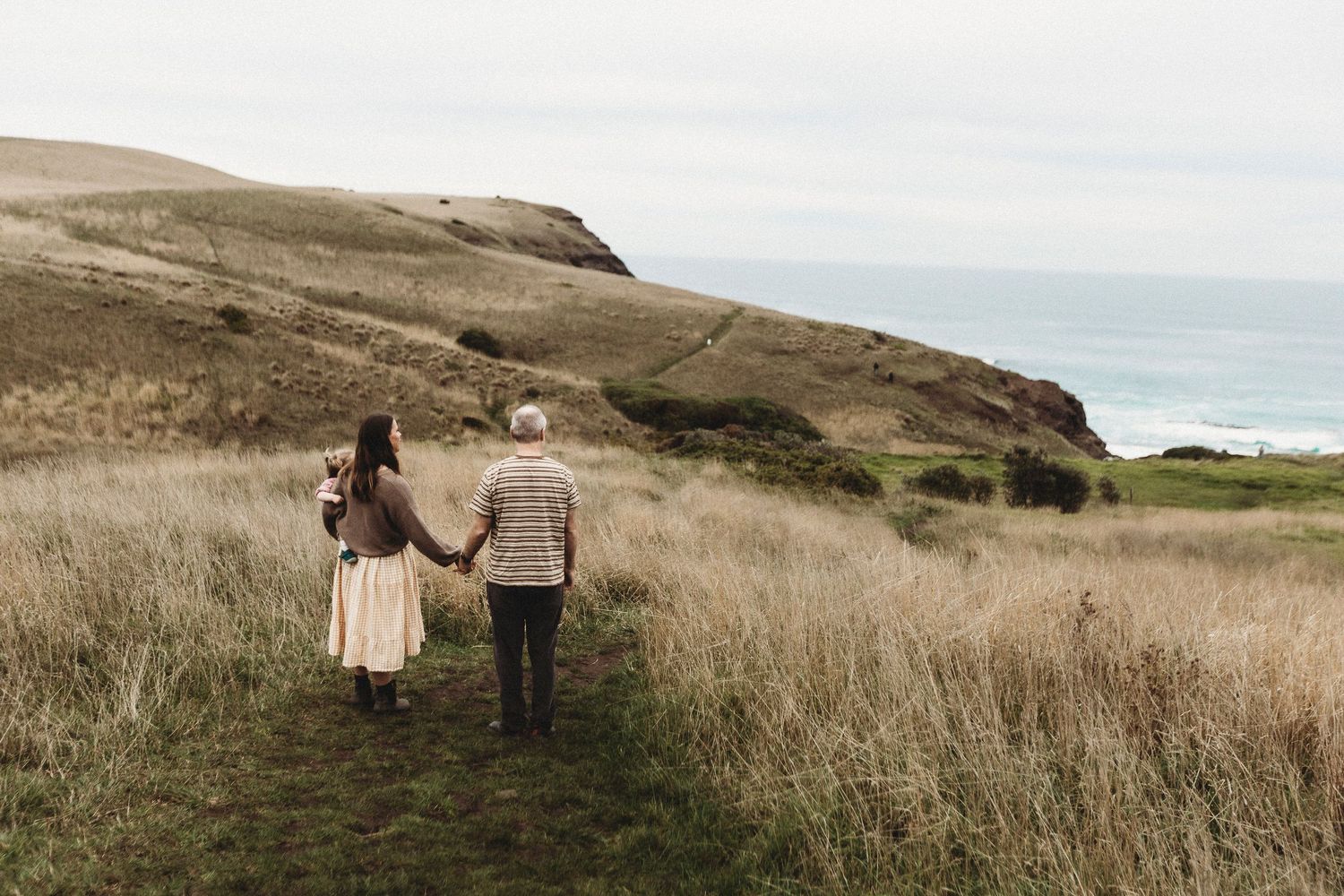 Couple walking hand in hand through golden coastal grasslands toward the ocean on a cloudy day.