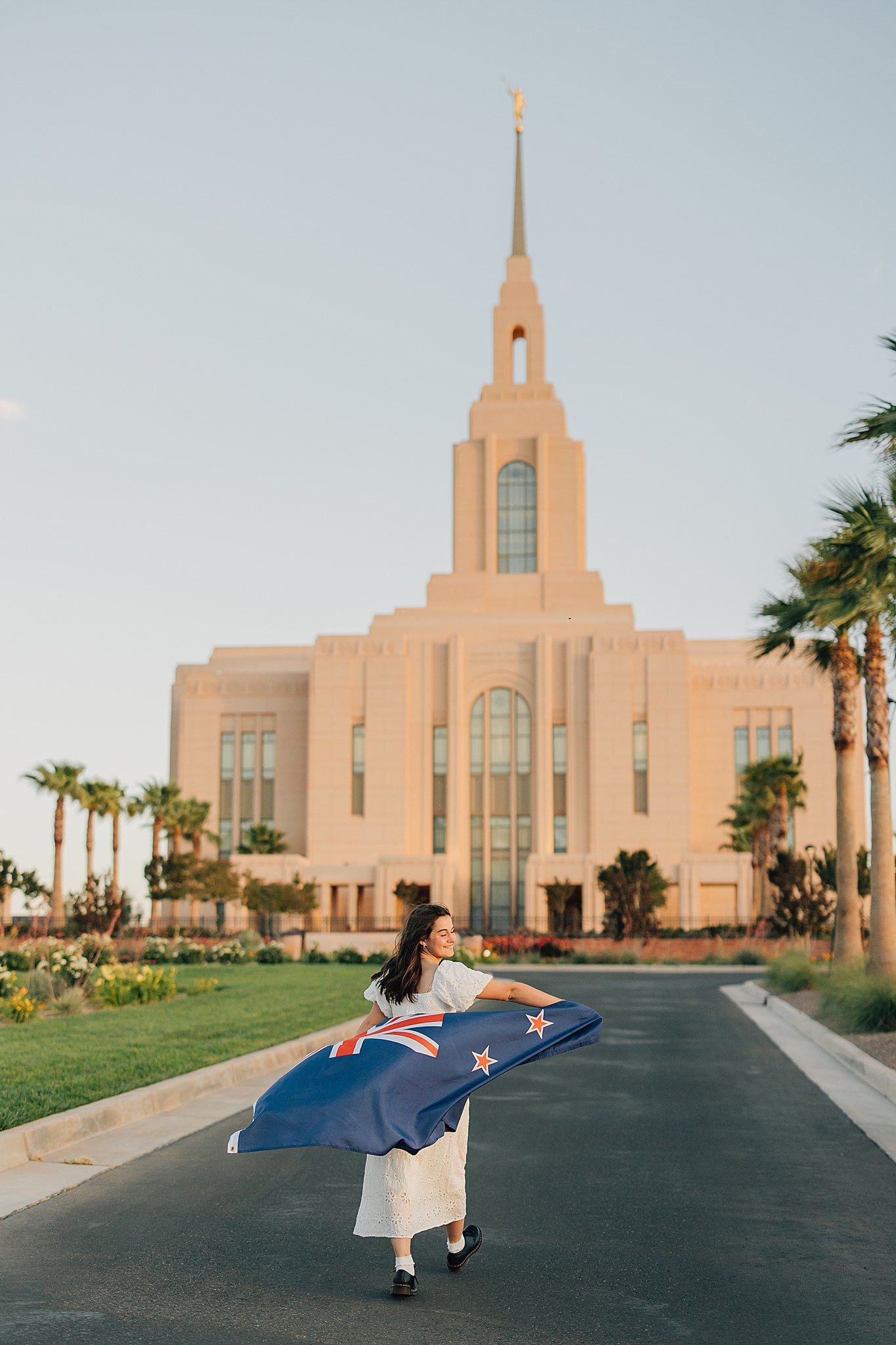 Woman holding New Zealand flag in front of an LDS temple with palm trees and clear sky.