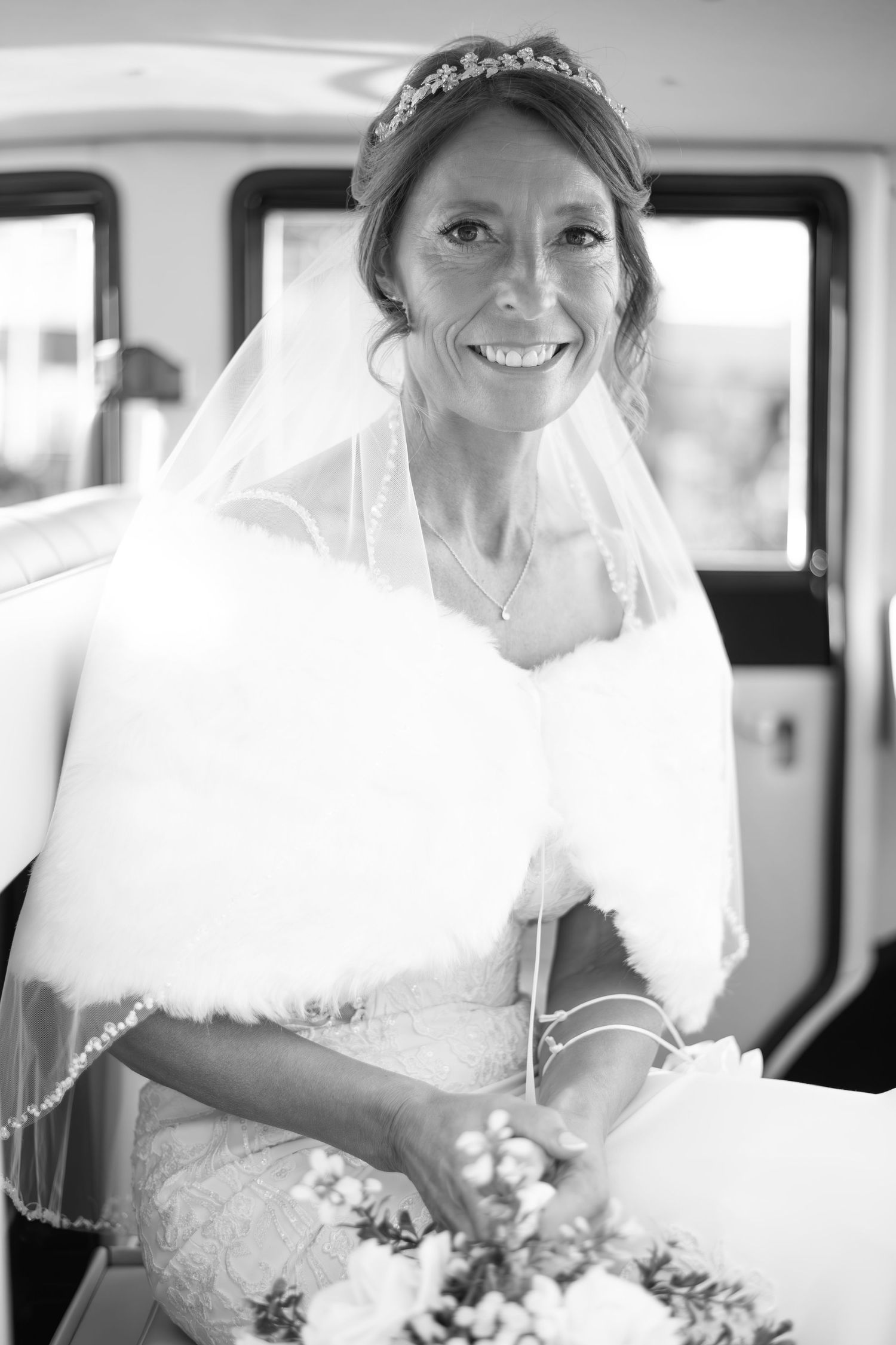 Smiling bride in veil, tiara, fur stole, holding bouquet, black and white wedding photo.