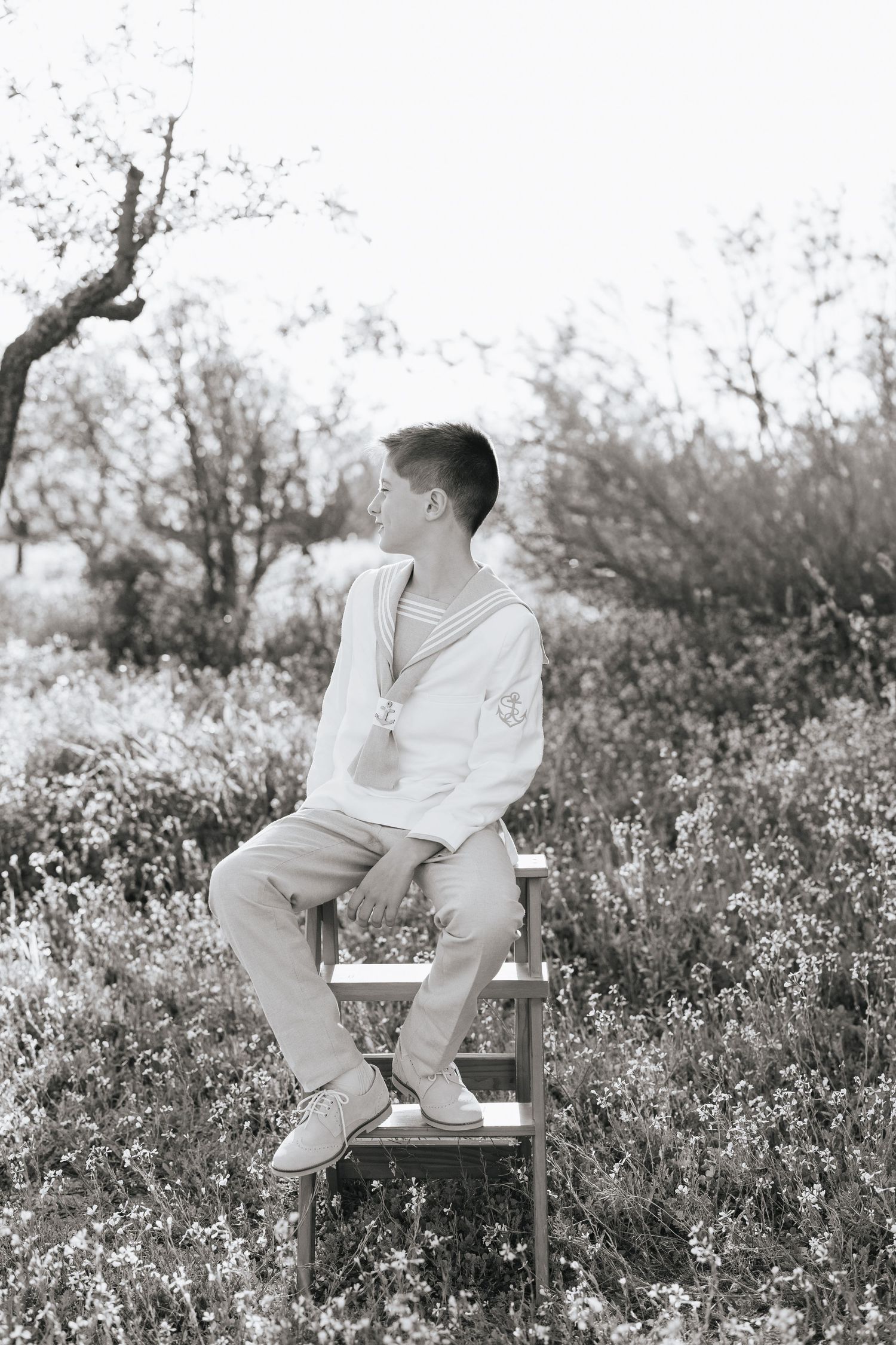 Black and white photo of a young man sitting on a wooden chair outdoors among wildflowers and trees.