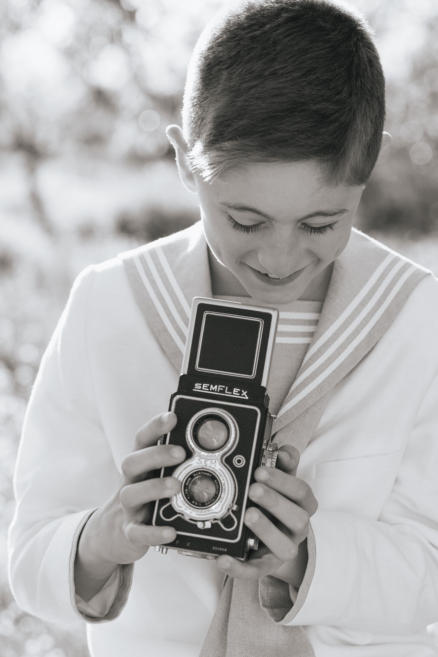 Black and white photo of a smiling boy in a sailor outfit holding a vintage Semflex twin-lens reflex camera outdoors.