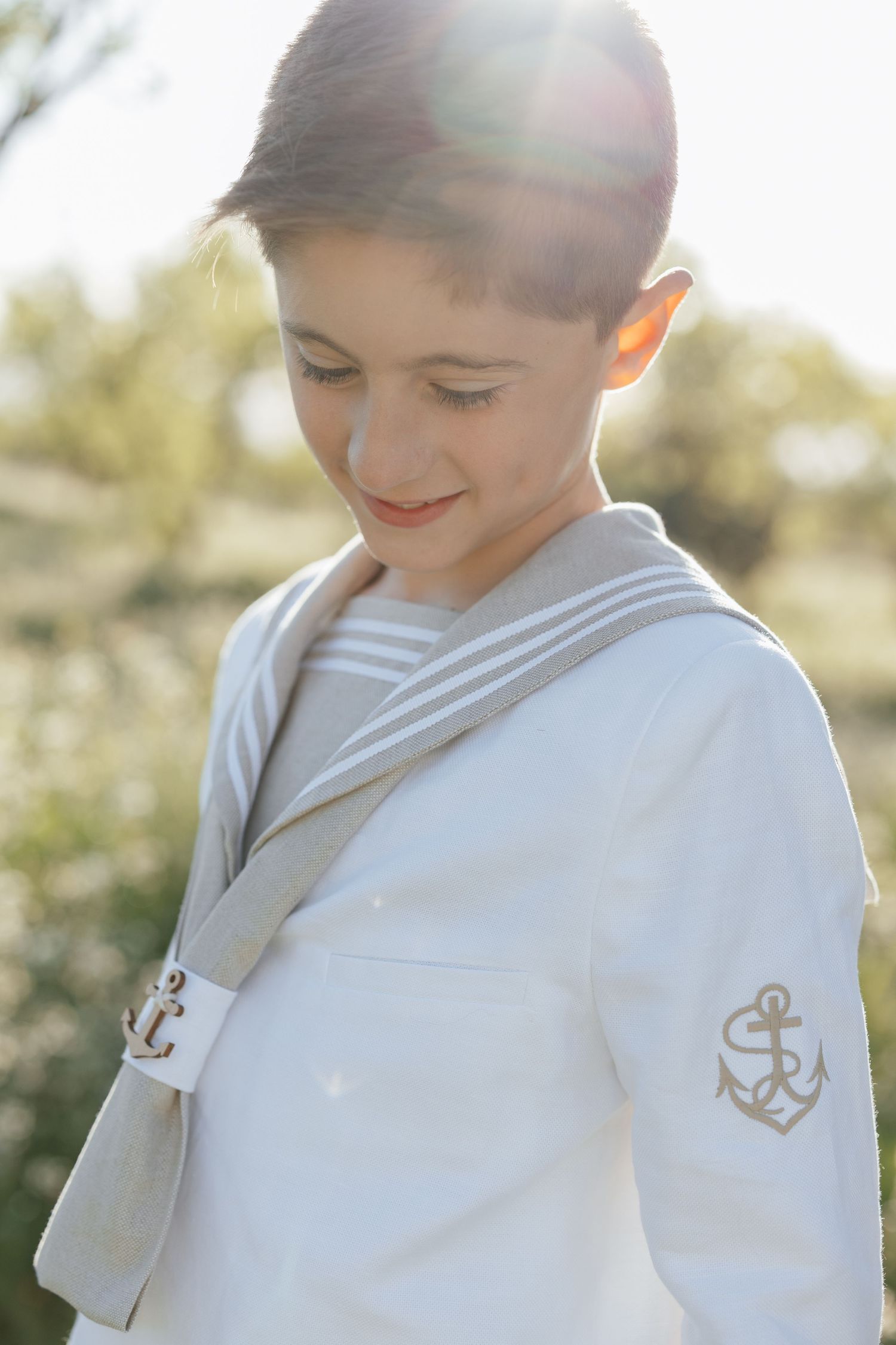 Young boy in white sailor outfit with anchor emblem, smiling outdoors in soft sunlight.