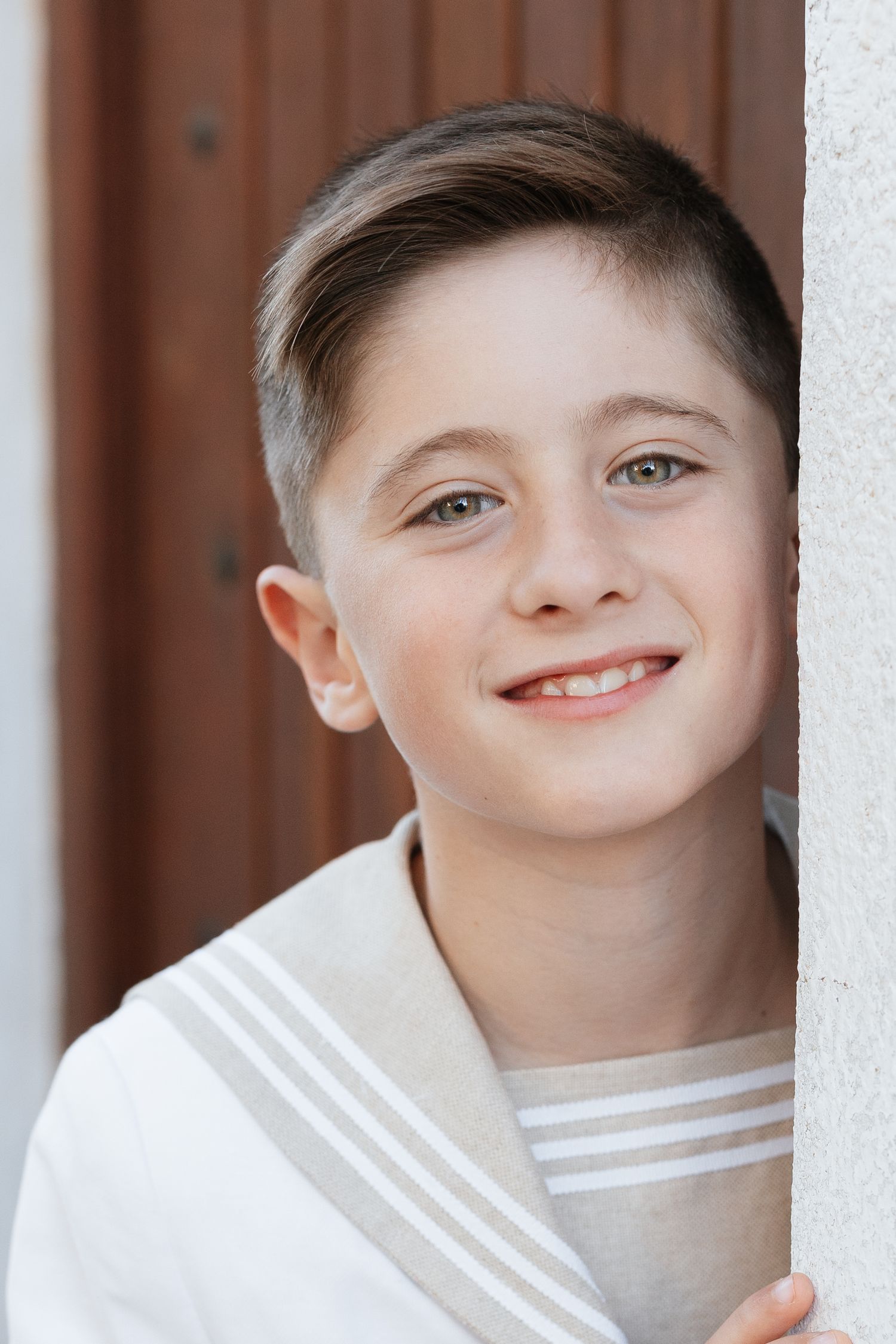 Smiling young boy with short brown hair leaning against a white wall, wearing a striped shirt outdoors.