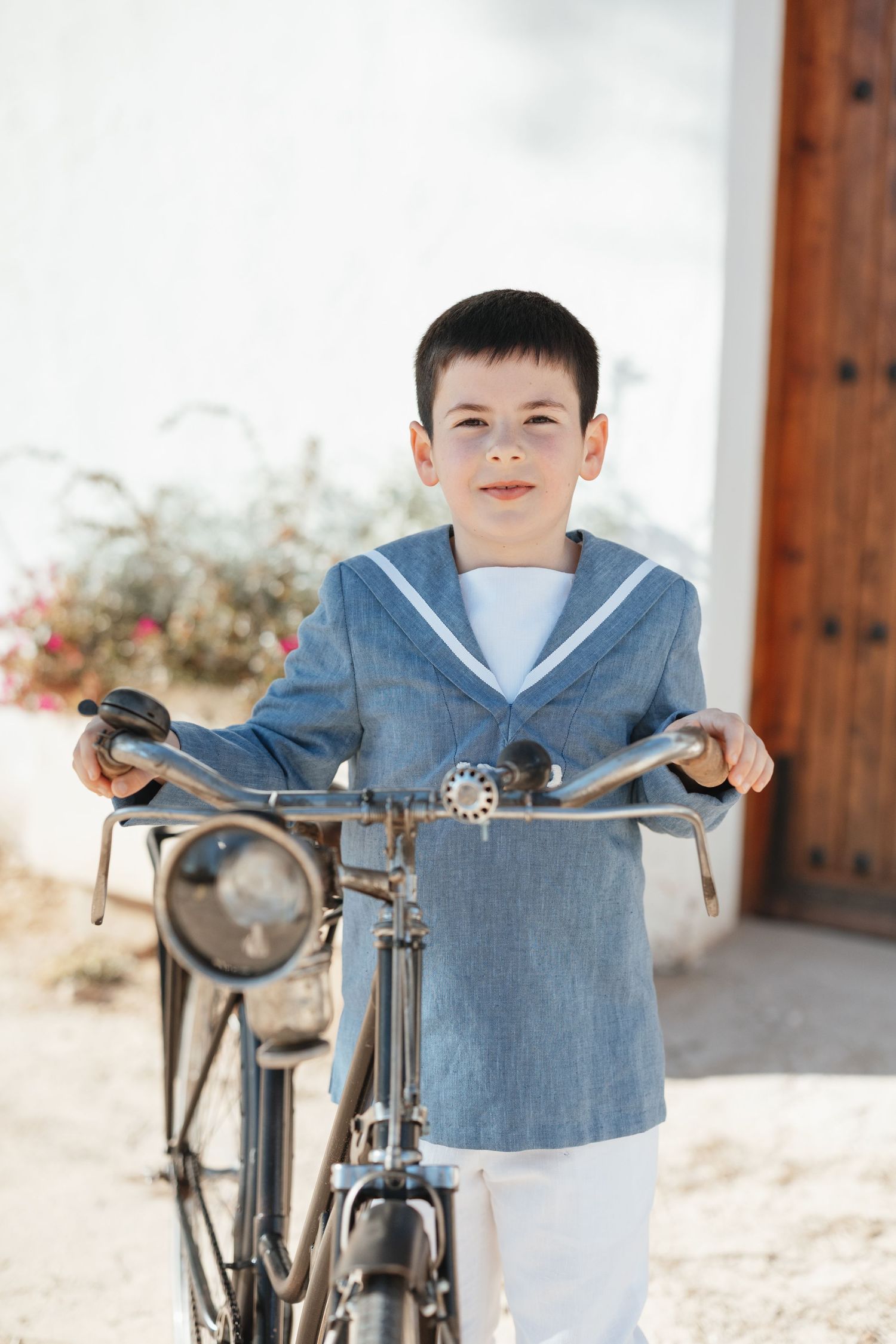 Young boy in sailor outfit stands with vintage bicycle outdoors on sunny day.