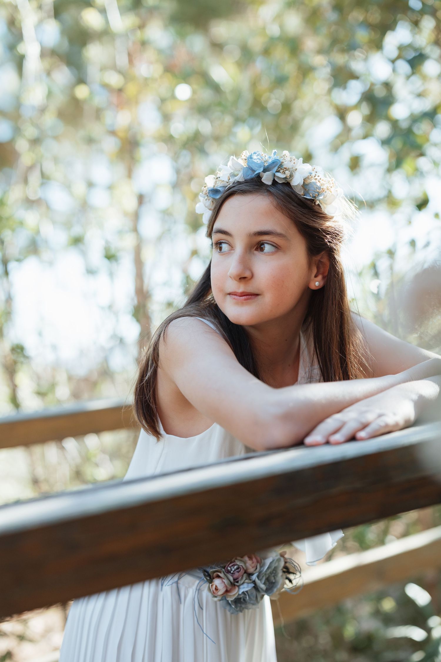 Young girl in white dress and blue floral crown leans on wooden fence, gazing dreamily at nature backdrop.