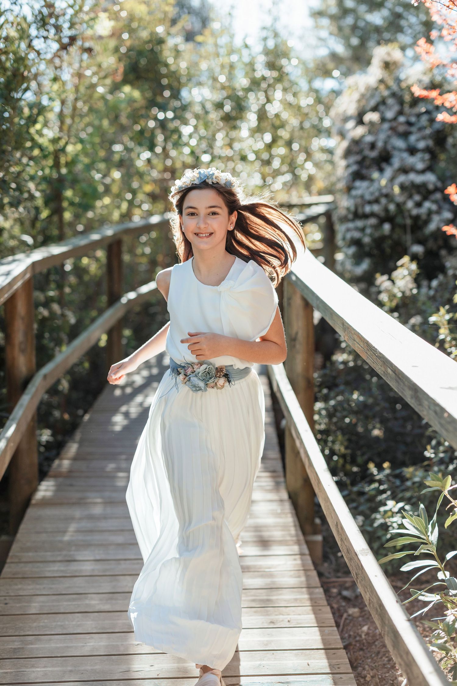 Happy girl in white dress with floral crown running on wooden bridge outdoors.