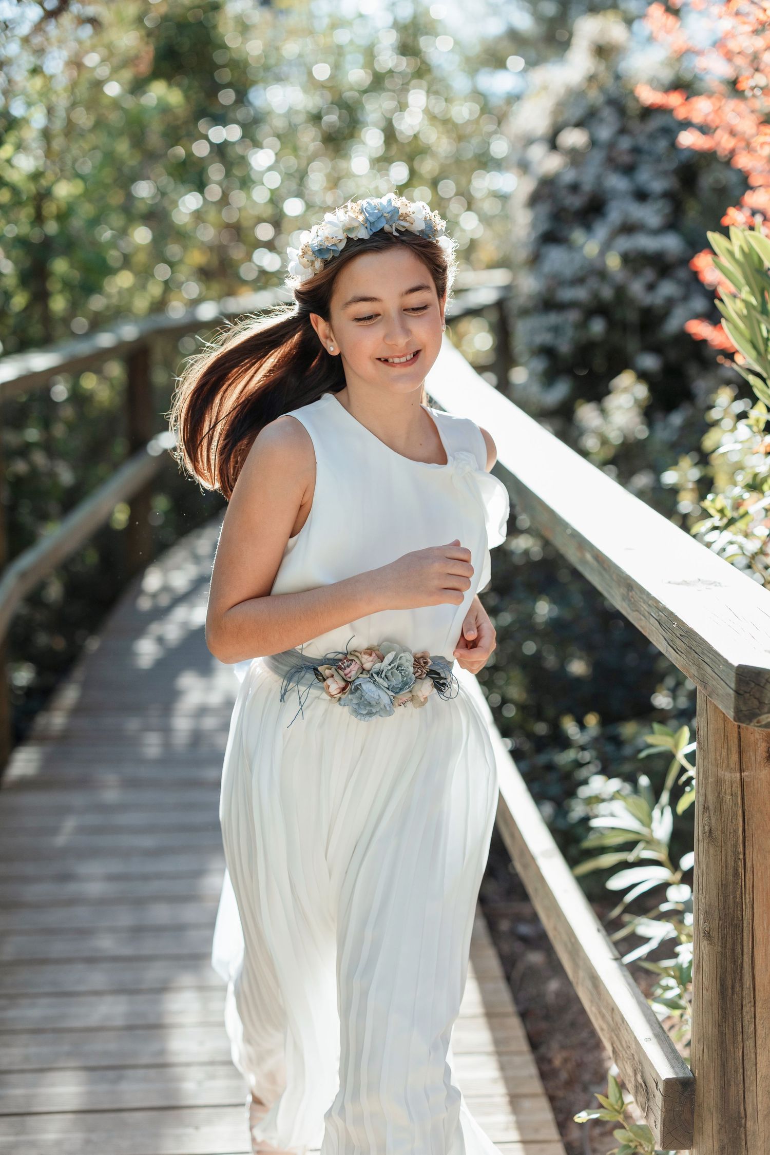 Young girl in white dress and floral crown walks on wooden bridge surrounded by lush green garden.