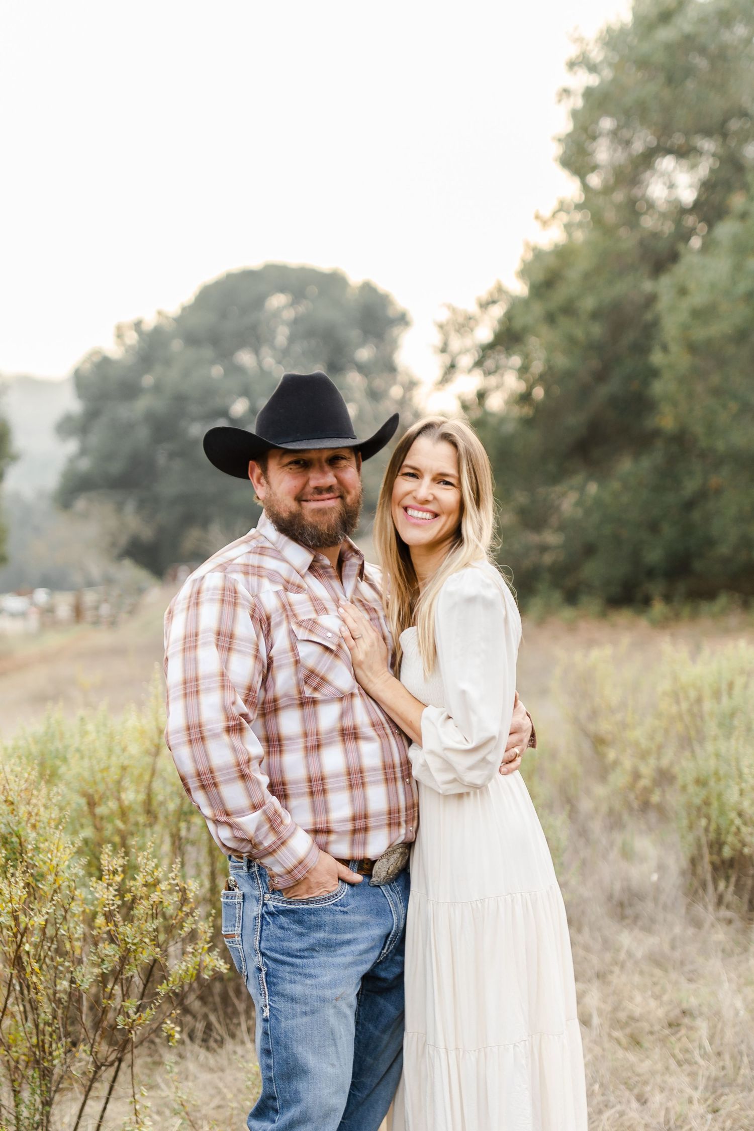 A smiling couple poses outdoors, man in cowboy hat and plaid shirt, woman in white dress, in a golden field.