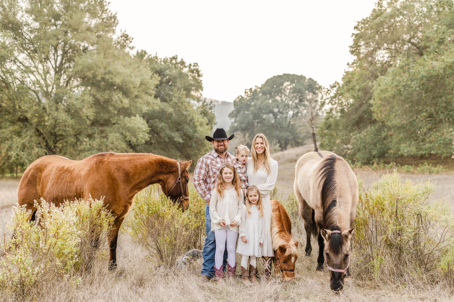 Family of five poses outdoors with horses grazing nearby in a scenic field surrounded by lush green trees.