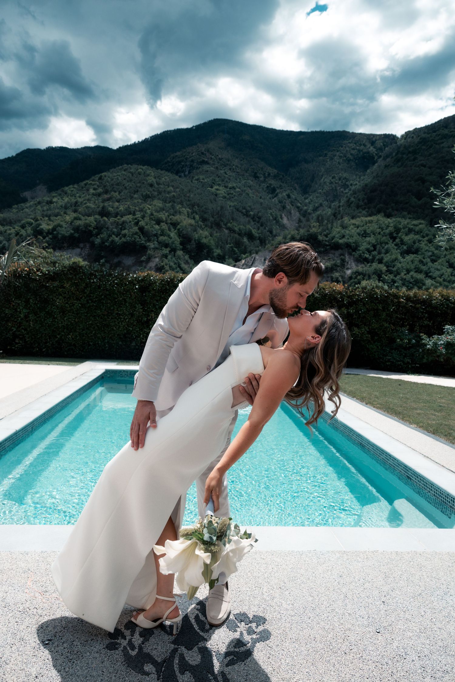 Bride and groom share romantic dip kiss by pool with mountain backdrop on wedding day.