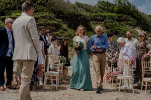 Bride in teal skirt walks down sandy beach aisle with escort toward waiting groom.