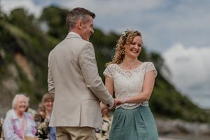 Smiling bride in lace top and teal skirt holds hands with groom in beige suit at beach wedding.