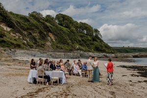 Wedding party seated at table on sandy beach with lush green hillside backdrop.