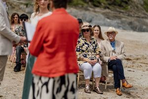 Wedding guests seated on a sunny beach watching a seaside ceremony with rocky cliffs behind.