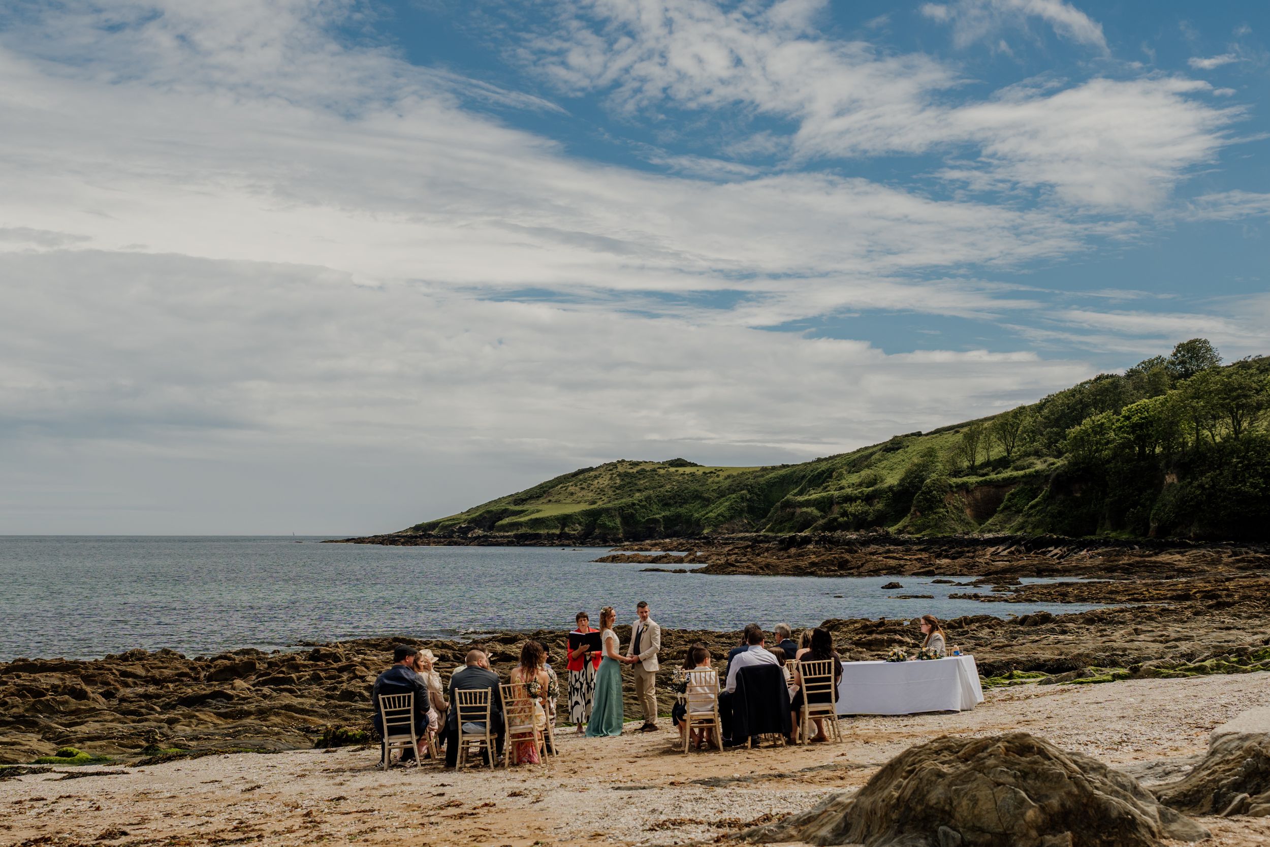 Small intimate beach wedding ceremony with guests seated facing the ocean under blue skies.
