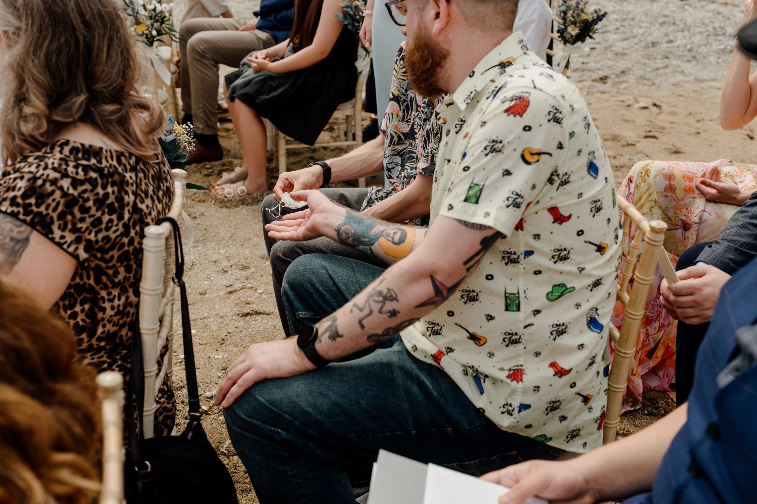 Tattooed wedding guest in printed shirt seated at an outdoor beach ceremony.