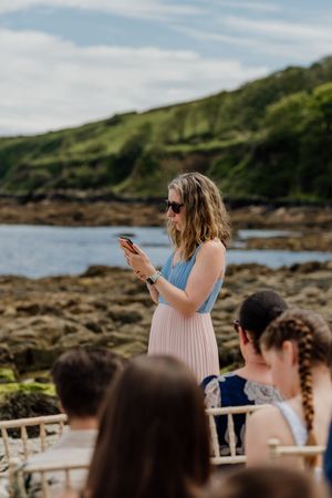 Wedding guest in pink pleated skirt photographs ceremony on rocky coastal shoreline.