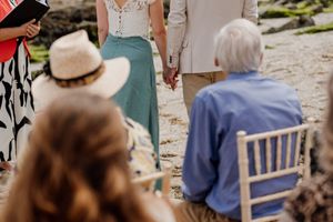 Couple holds hands during beach wedding ceremony while guests watch from white chairs.