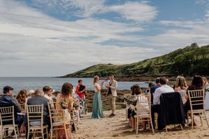 Outdoor beach wedding ceremony with guests seated on chairs facing couple at shoreline.