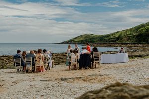 Outdoor beach wedding ceremony with guests seated on chairs by rocky shoreline with green hills and sea backdrop.