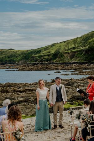 Smiling bride and groom hold hands at beach wedding ceremony with rocky coastline behind them.