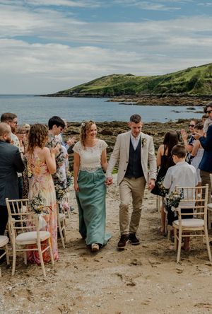 Newlywed couple walks hand-in-hand down sandy beach aisle as guests applaud during outdoor coastal wedding ceremony.
