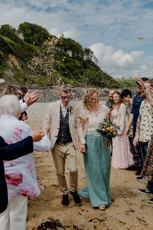 Bride and groom walk through confetti shower on sandy beach with lush green cliffs during seaside wedding celebration.