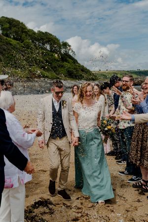 Newlyweds walk through confetti shower on sandy beach surrounded by cheering wedding guests.