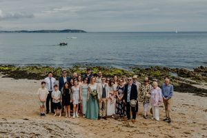 Large wedding party group photo on a rocky beach with ocean and distant headland in the background.