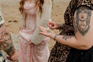 Tattooed hands exchanging small items on sandy beach during outdoor bohemian wedding ceremony.