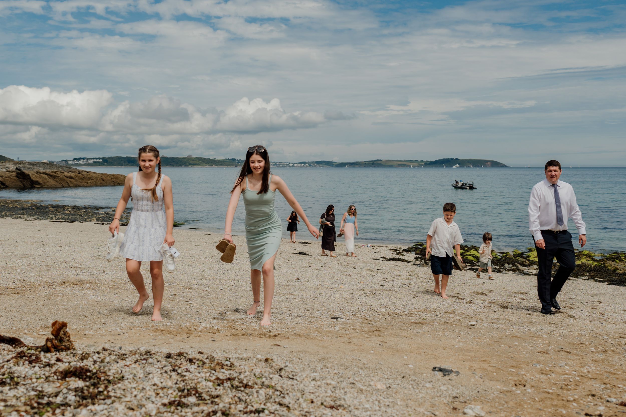 Wedding guests walking barefoot along a pebble beach with a scenic ocean backdrop.