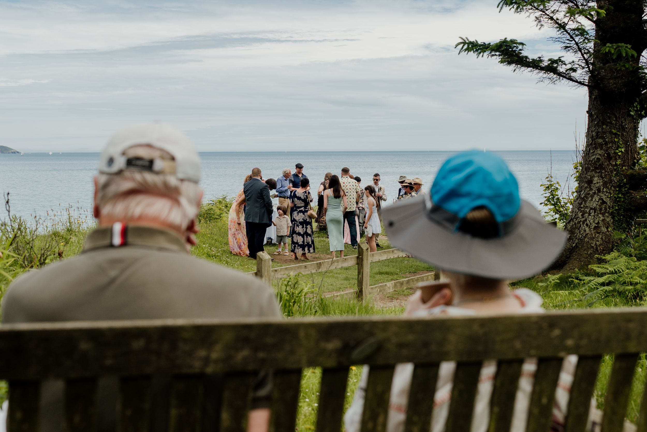 Two elderly guests sit on a bench overlooking a seaside wedding ceremony with ocean views.