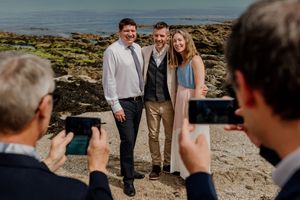 Groom posing with two guests on a rocky beach while attendees photograph the moment.