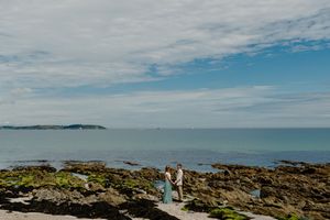 Bride and groom standing on rocky shoreline with vast blue ocean and cloudy sky behind them.