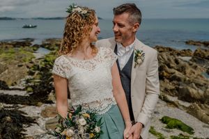 Smiling bride and groom hold hands on rocky coastline with blue ocean in background.