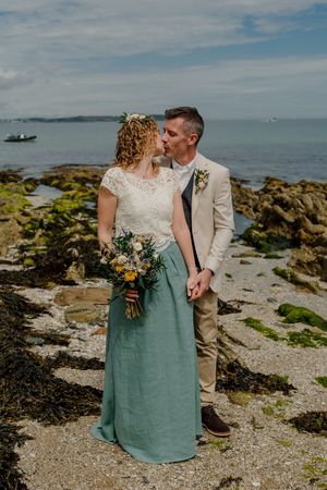 Newlyweds kiss on rocky beach, bride in lace top and teal skirt holding wildflower bouquet.
