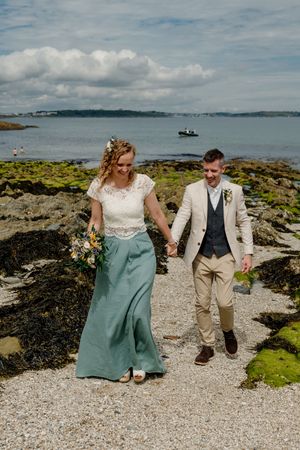 Bride in teal skirt and groom in beige suit walk hand in hand on a pebble beach by the sea.