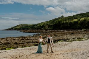Bride and groom hold hands walking on a sandy beach with dramatic green coastal cliffs behind them.