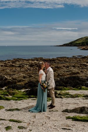 Bride in teal skirt and groom in tan suit share a romantic moment on a rocky Irish coastline under blue skies.
