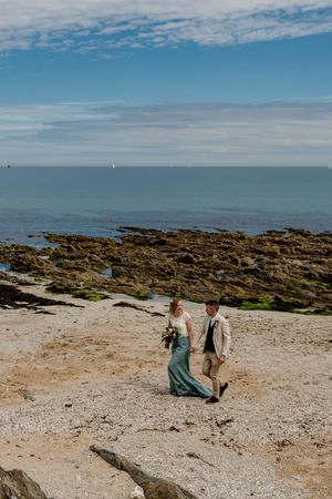 Bride in teal gown and groom in beige suit walk along a rocky coastal beach.