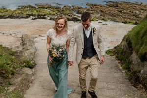 Smiling bride and groom walk hand-in-hand up seaside steps with a bouquet.
