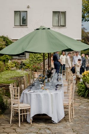 Elegant outdoor wedding reception table set with white linen and green umbrella in garden venue.