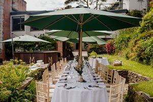Elegant outdoor dining setup with green umbrellas, white tablecloths, and wooden chairs in a lush garden setting.