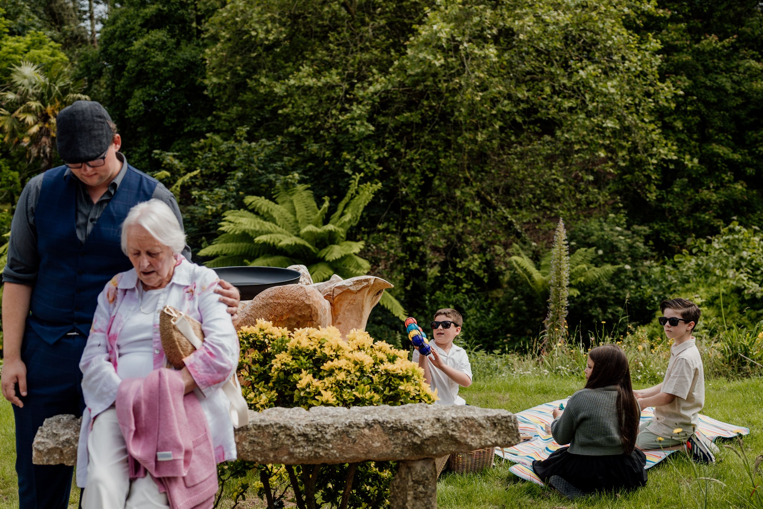 Elderly woman fans herself beside stone planter with yellow flowers at garden wedding.