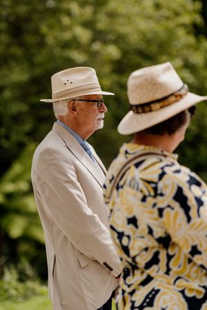 Two elderly wedding guests in straw hats and formal attire stand outdoors in green garden.