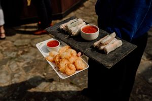A server holds a slate tray with spring rolls, fried shrimp, and two dipping sauces at an outdoor event.