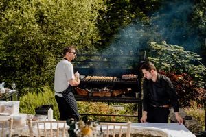 Two chefs grilling skewers on a large outdoor BBQ grill surrounded by lush greenery at an outdoor event.