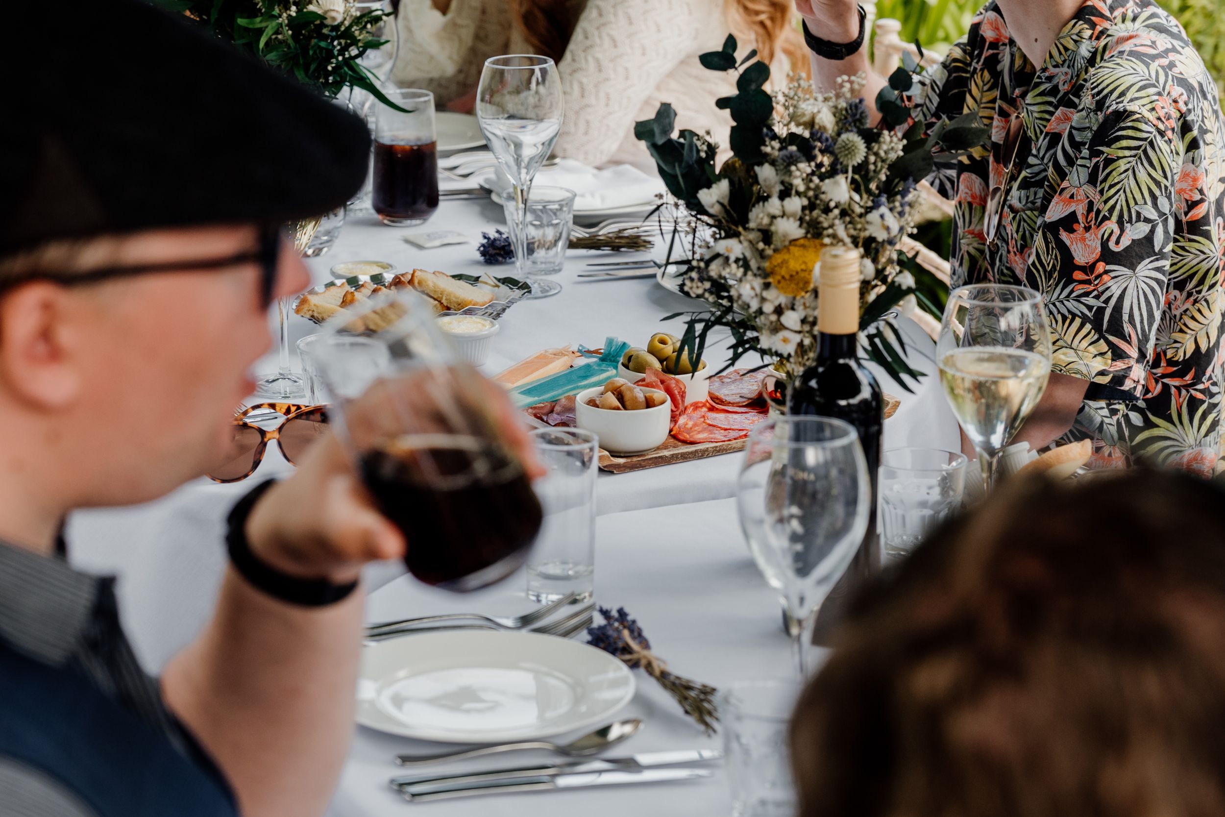 Guests enjoying a formal outdoor dinner party with wine, charcuterie, and floral centerpieces on a white tablecloth.